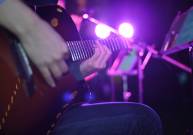 A person sits on a stool and plays their acoustic guitar on a dive bar stage